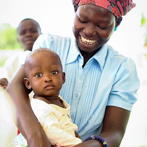 A woman in a headwrap holds a young child smiling as she looks at him. The child looks at the camera.